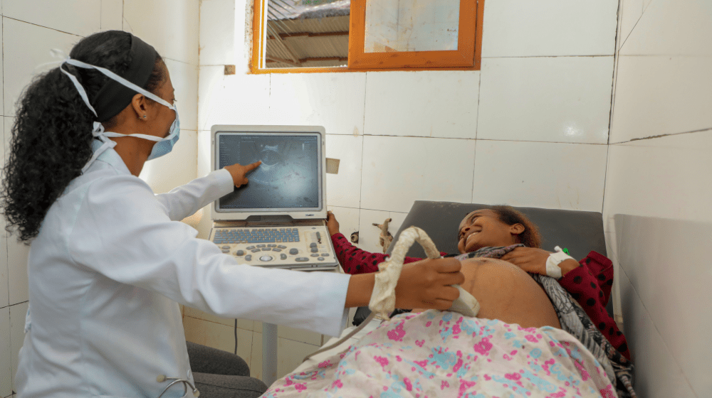 A health worker in a white medical coat and blue face mask gives an ultrasound to a smiling pregnant woman on a bed